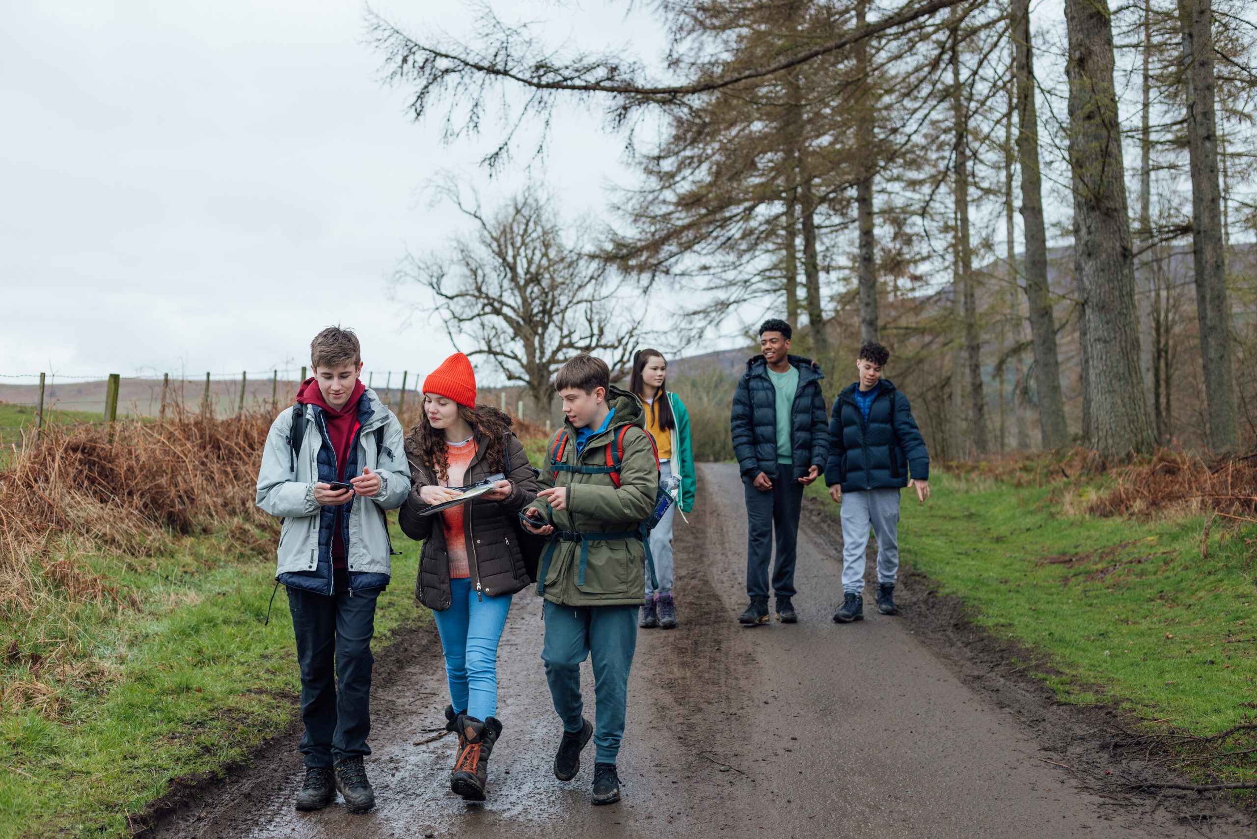 En grupp ungdomar i varma jackor vandrar tillsammans på en grusväg genom ett landskap med kala träd och gröna fält. Tre av dem i förgrunden studerar en karta och en mobiltelefon, medan de andra går bakom dem längs vägen, troligen under en friluftsdag eller orienteringsövning.