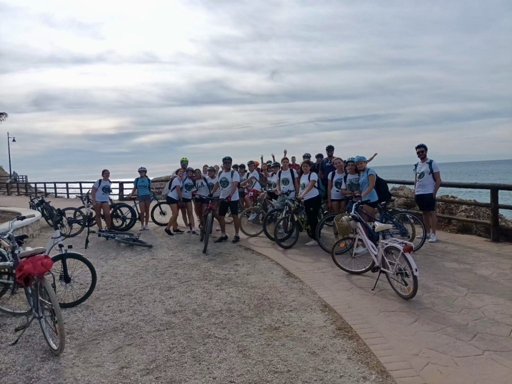 En stor grupp personer i vita t-shirts står med sina cyklar på en strandpromenad vid havet under en mulen dag, troligen efter en gemensam cykeltur.