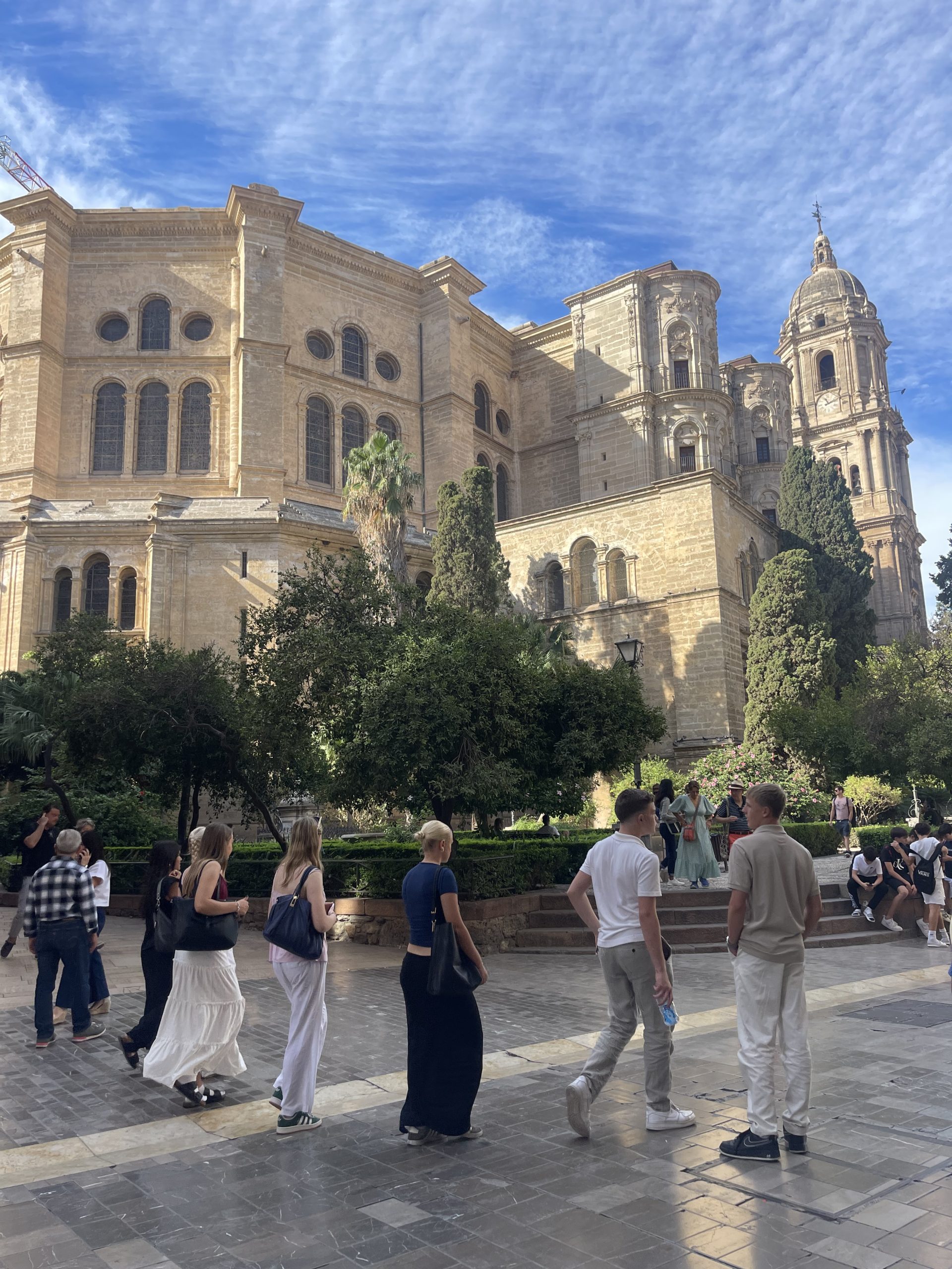Besökare promenerar och samlas på ett torg framför en stor stenkatedral med torn och dekorerad fasad, med palmer och grönska i förgrunden, troligen fotograferat under en resa.