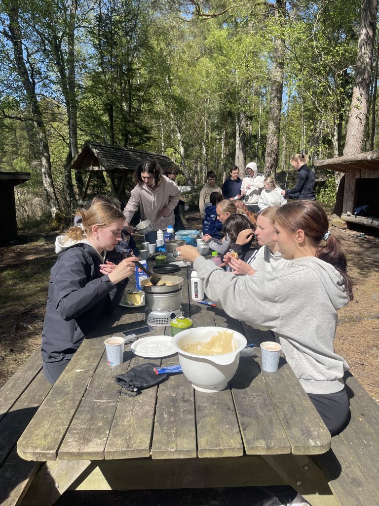 En grupp ungdomar sitter i skogen med matsäck vid ett träbord.