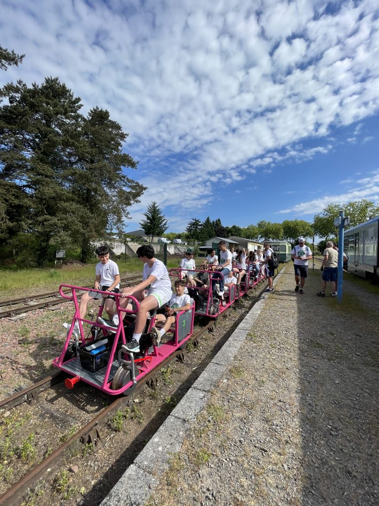 En grupp barn och vuxna åker på en rosa trampdressin på räls genom ett grönskande landskap en solig dag med blå himmel.