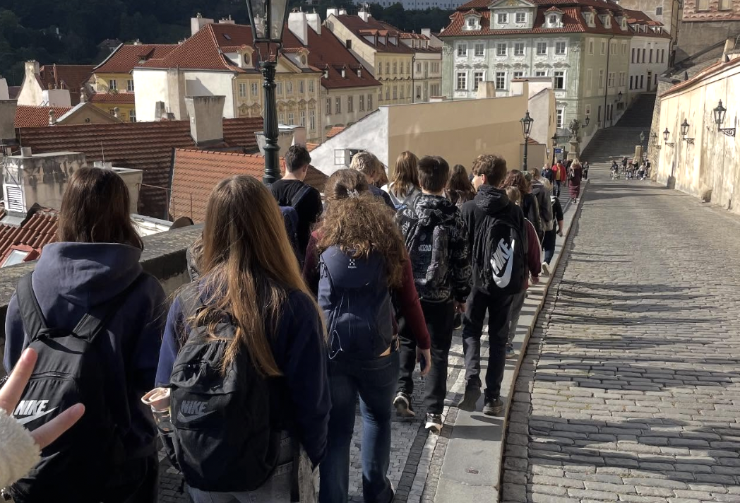 En grupp ungdomar med ryggsäckar går längs en kullerstensgata i en europeisk stad med gamla byggnader med rödtegel tak, troligen under en skolresa.
