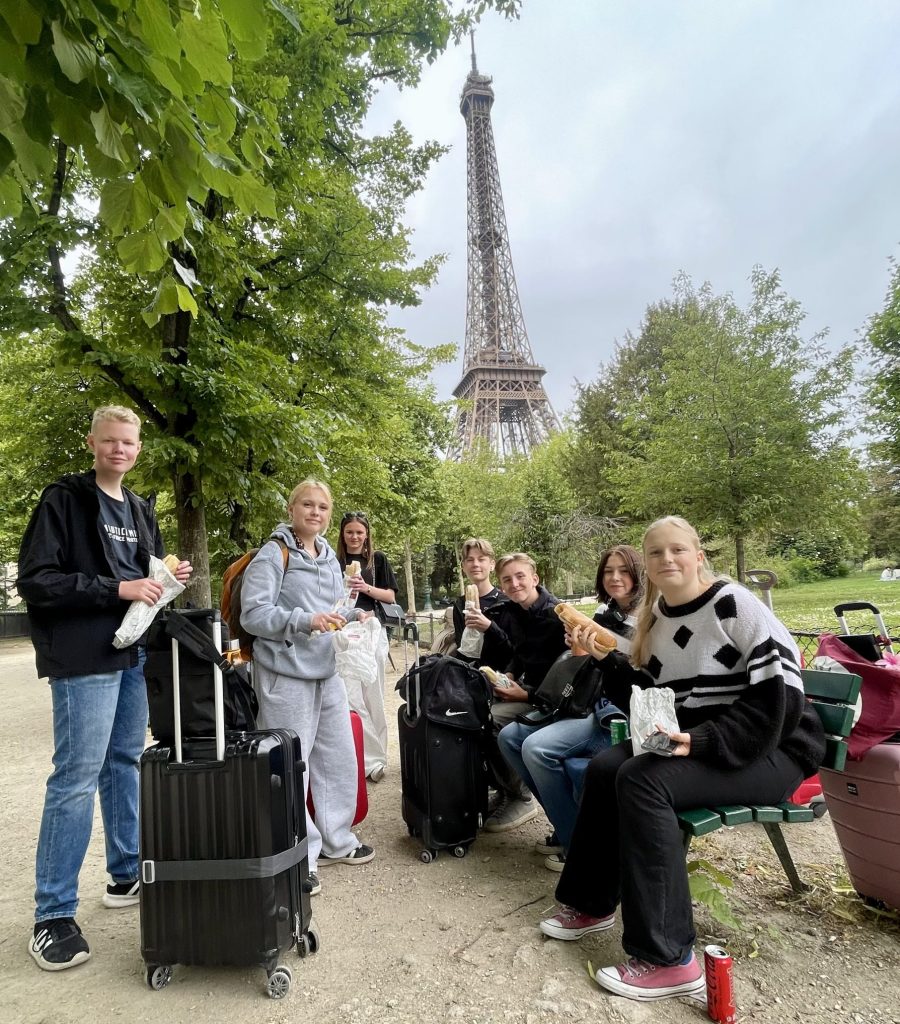En grupp ungdomar med resväskor poserar tillsammans utomhus i en park med Eiffeltornet synligt i bakgrunden, troligen under en skolresa till Paris.