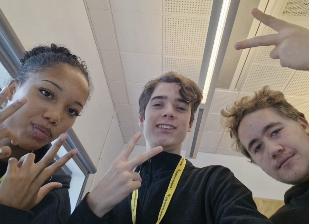 Selfie tagen underifrån av tre ungdomar i ett klassrum med vitt undertak och lysrörsarmatur. Alla tre gör peace-tecken med händerna och ler eller poserar mot kameran. En av dem bär ett gult nyckelband.