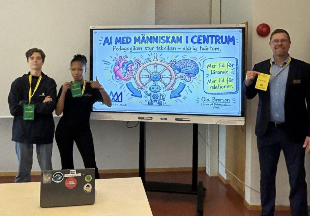 Två elever och en man i kavaj håller upp sina namnbrickor bredvid en skärm. På skärmen står “AI med människan i centrum – Pedagogiken styr tekniken, aldrig tvärtom”, med en tecknad bild av ett hjärta och en hjärna vid ett skeppsratt.