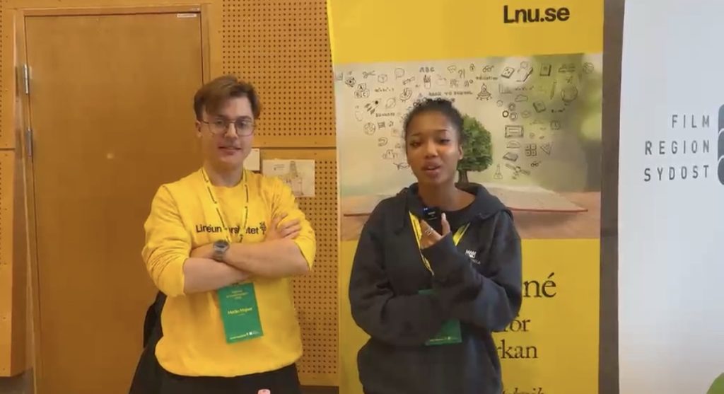 En ung man i gul Linnéuniversitetet-tröja står med armarna i kors bredvid en elev som pratar i en liten mikrofon. Bakom dem syns en gul roll-up från Lnu.se och en banderoll från Film Region Sydost.