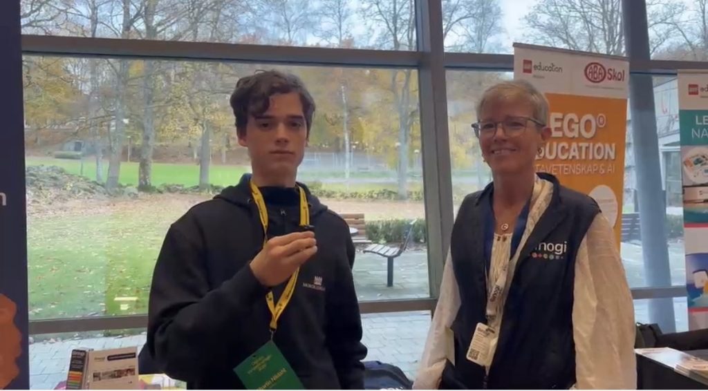 En elev med mikrofon intervjuar en kvinna i väst och glasögon vid en utställningsmonter. I bakgrunden syns roll-ups från LEGO Education (“Datavetenskap & AI”) och ABA Skol, och genom stora fönster en park med höstlöv.