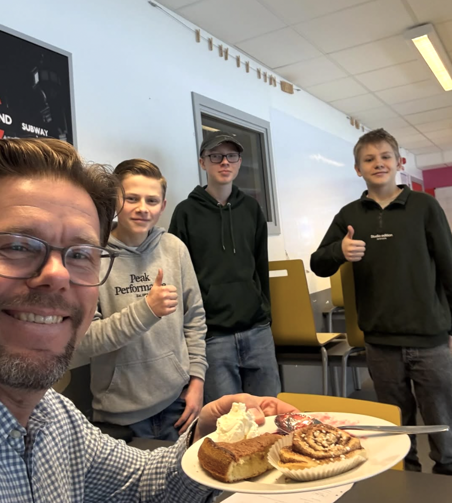 En leende man med glasögon tar en selfie medan han håller upp en tallrik med äppelkaka, vispgrädde och kanelbulle. Bakom honom står tre tonårskillar i ett klassrum, två av dem gör tummen upp.